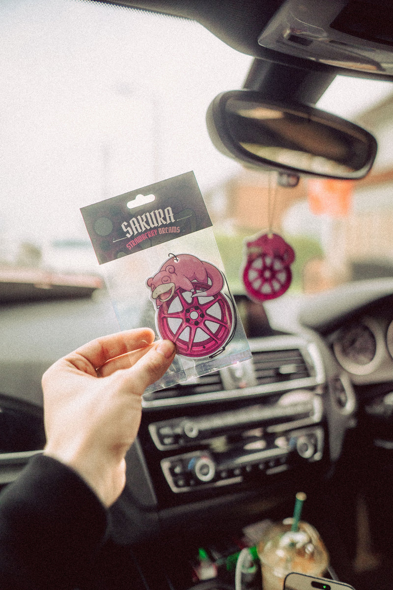 Hand holding a Sakura car air freshener package in a car interior.