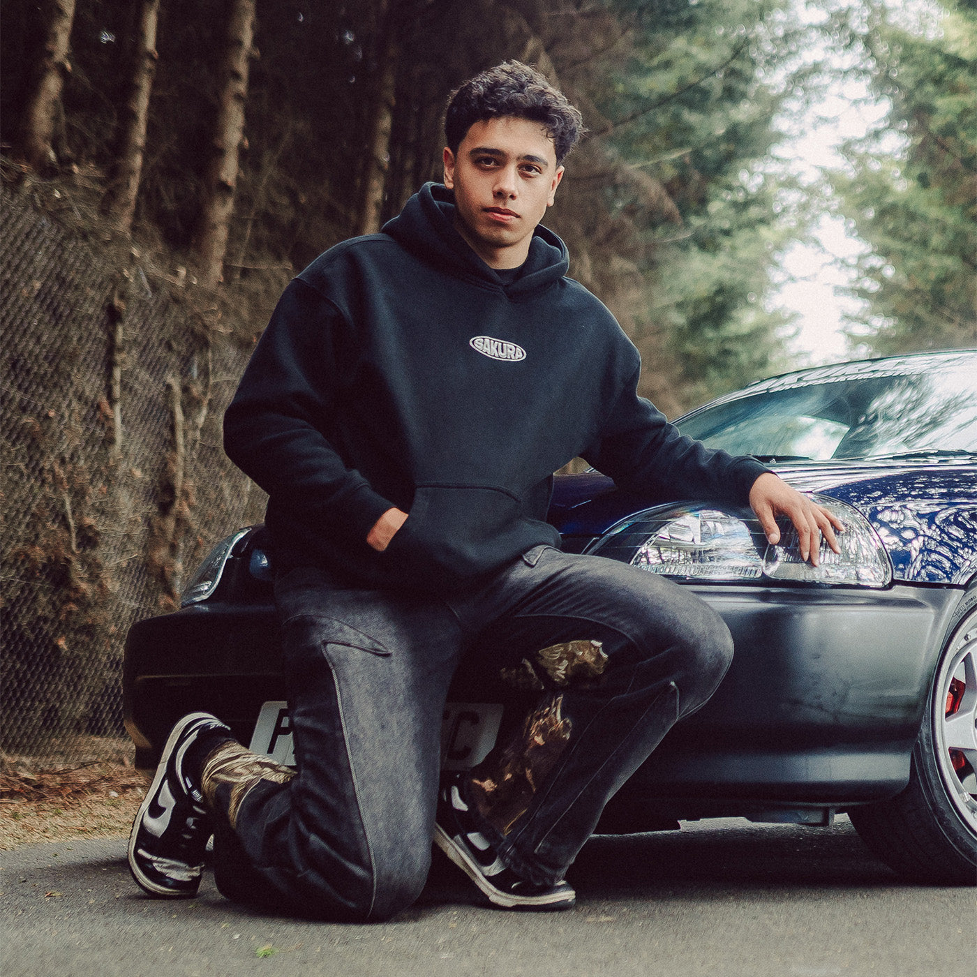 A young man wearing a black hoodie with a small chrome sakura chest logo, dark gray jeans, and black-and-white sneakers, kneeling on the front bumper of a honda civic ek9 in an outdoor wooded setting.