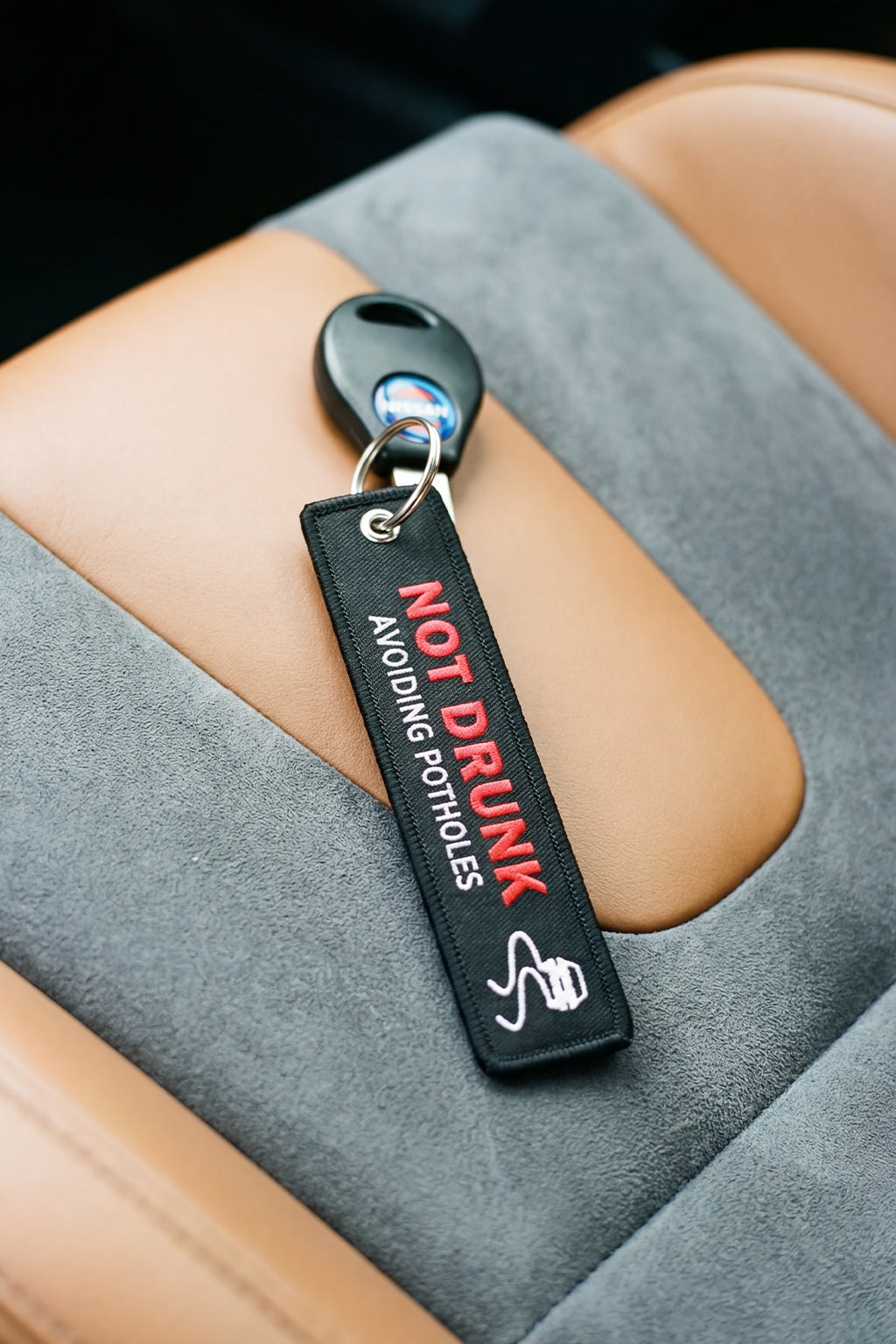 Keychain with 'Not Drunk Avoiding Potholes' text on a car seat.