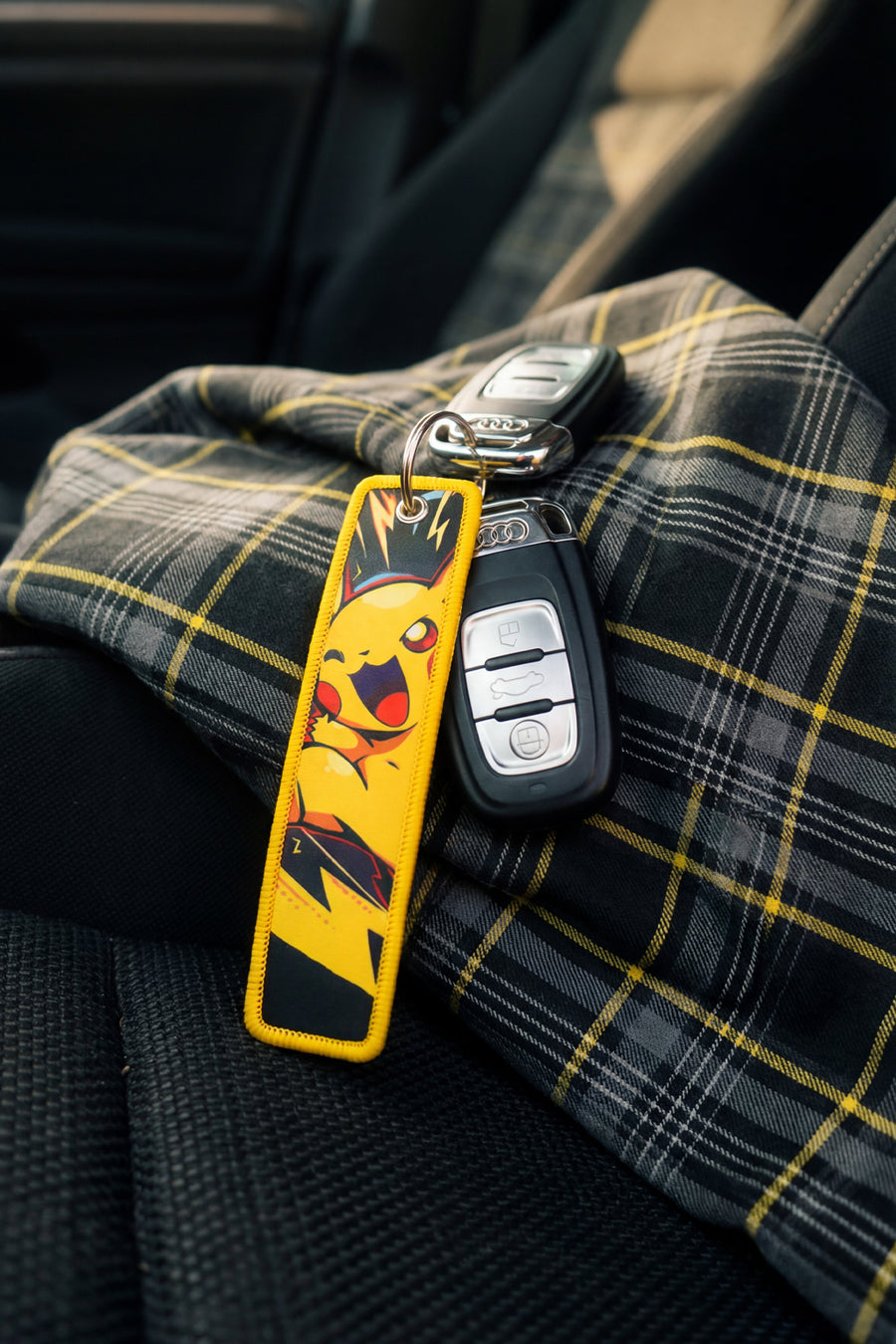 Keychain with a Pika character attached to a car key on a tartan fabric surface inside a vehicle.