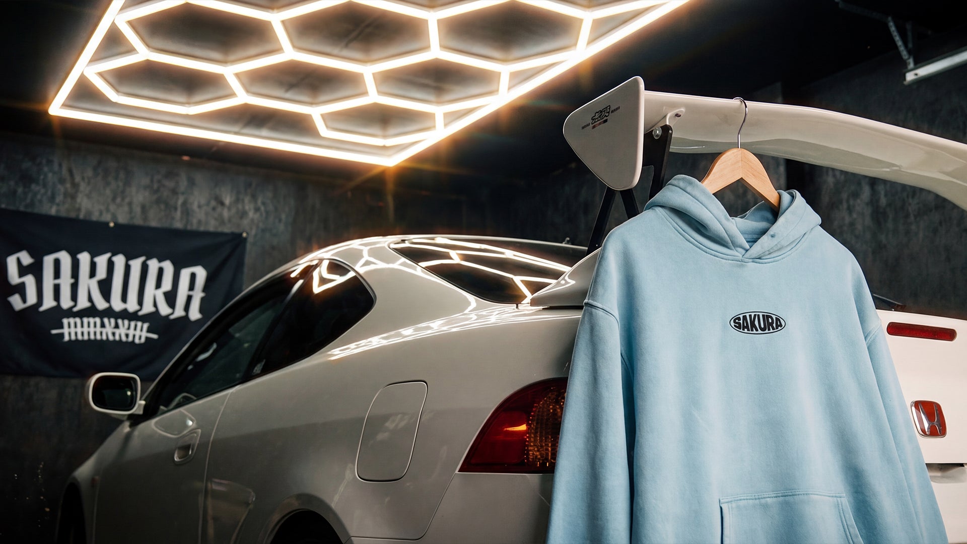 Light blue hoodie with 'Sakura' logo hanging on a car in a garage setting.