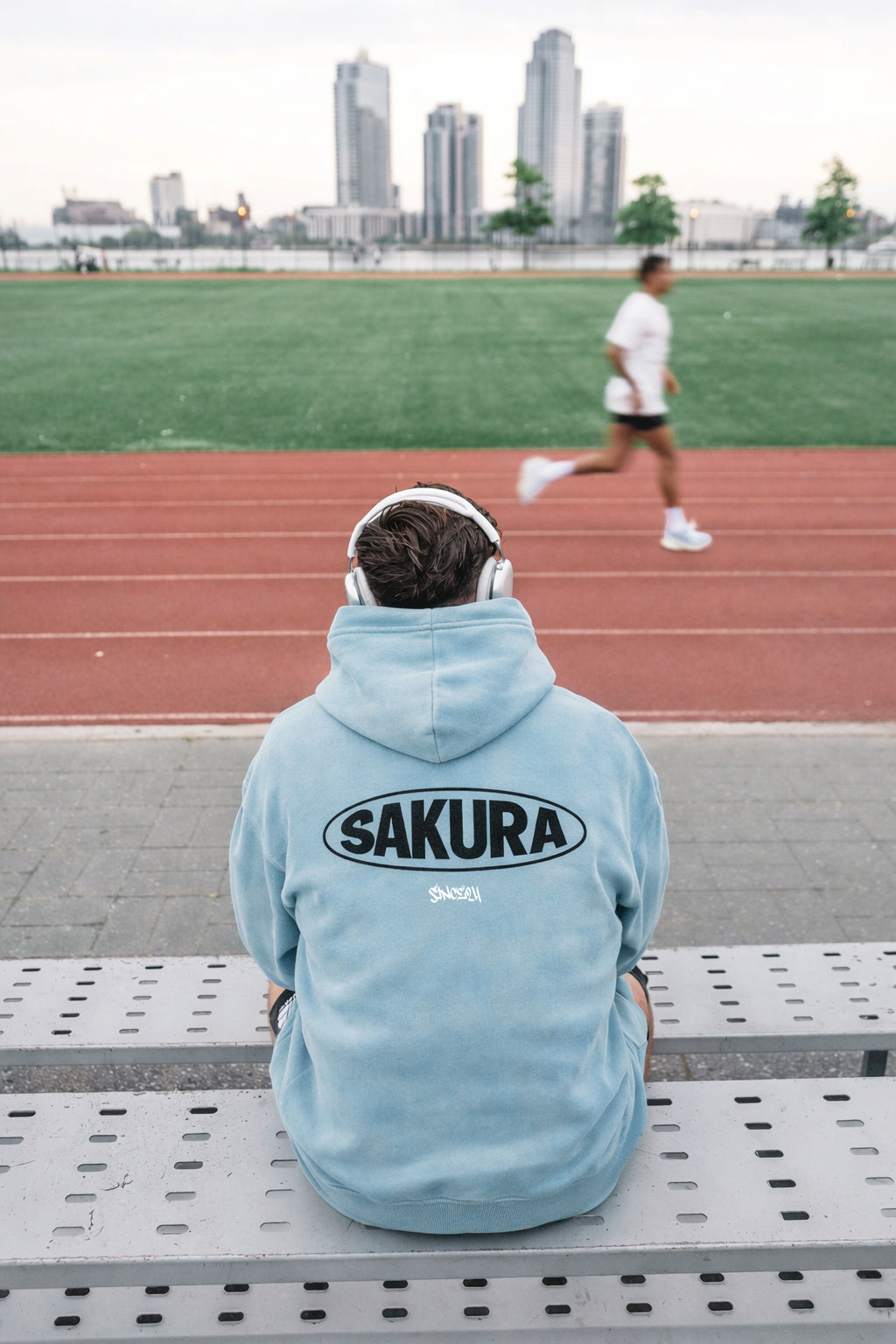 Person wearing a light blue hoodie with 'Sakura' branding, sitting on a bench in an outdoor setting with a runner in the background.