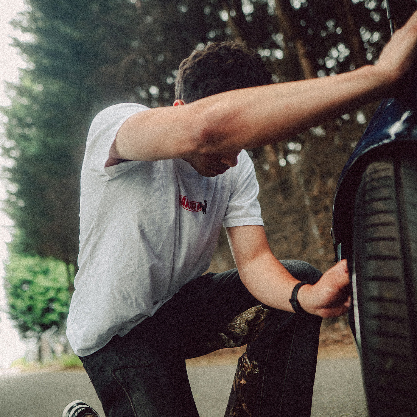 A man in a white t-shirt and dark jeans crouches beside a car tire outdoors, with trees in the background. The t-shirt features a small red graphic on the chest.